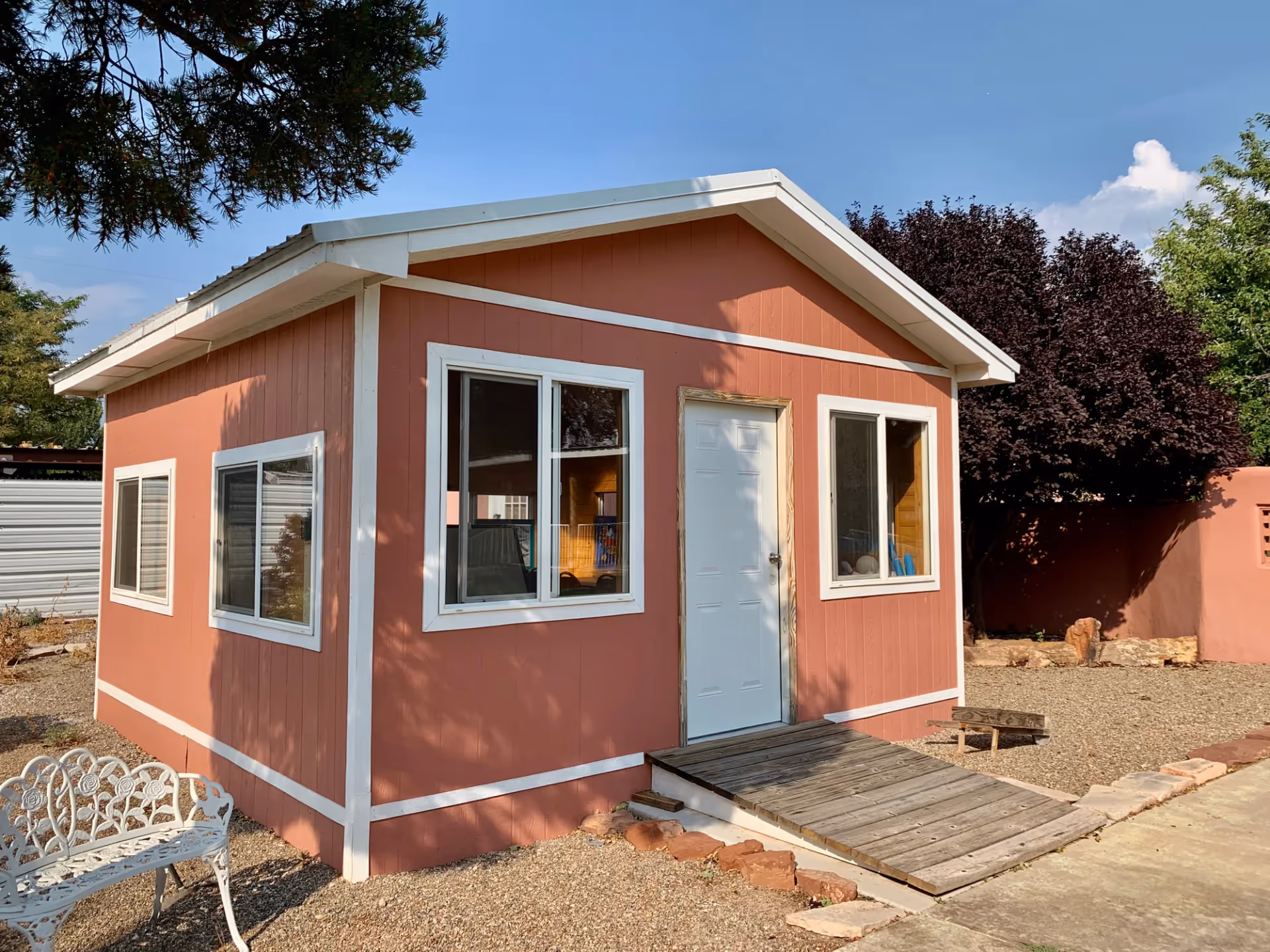 Small single-story building painted in reddish-brown with white trim around windows and door, featuring a wooden ramp leading to a white door. The building is surrounded by gravel and some trees, with a white metal bench on the left side.