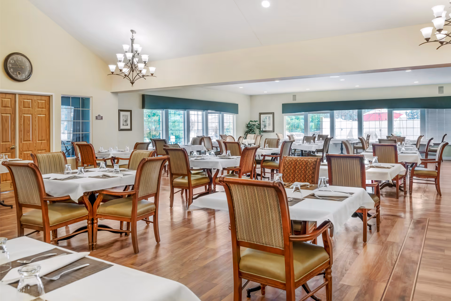 A spacious dining room with multiple tables covered in white tablecloths, each set with napkins and upside-down glasses. The room has wooden floors, large windows with green valances letting in natural light, and chandeliers hanging from the ceiling. There are wooden chairs with cushioned seats around the tables.