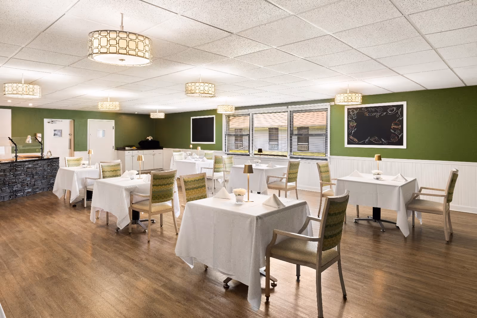 A bright dining room with multiple tables covered in white tablecloths, each set with napkins and small lamps. The room has green walls, large windows letting in natural light, wooden flooring, and modern ceiling lights. There is a stone-faced counter on the left side and a chalkboard on the right wall.