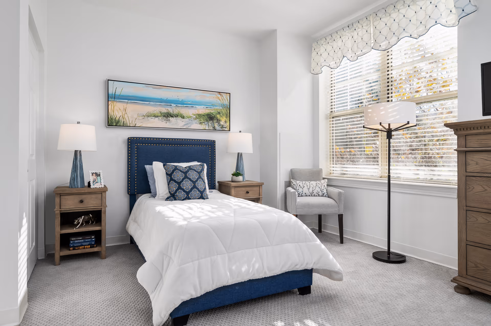 Bright, neatly furnished single bedroom with a blue upholstered twin bed, nightstands and lamps, an armchair, and a large window with blinds.