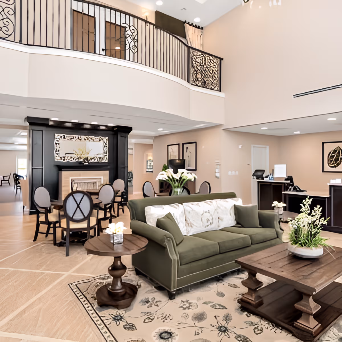 Spacious and well-lit assisted living facility common area featuring a green sofa with decorative pillows, wooden coffee and side tables with flower arrangements, a patterned area rug, and a fireplace with a large decorative mirror above it. There are several round tables with chairs around the room, framed artwork on the walls, and a reception desk in the background. The area has a high ceiling with a balcony railing visible above.