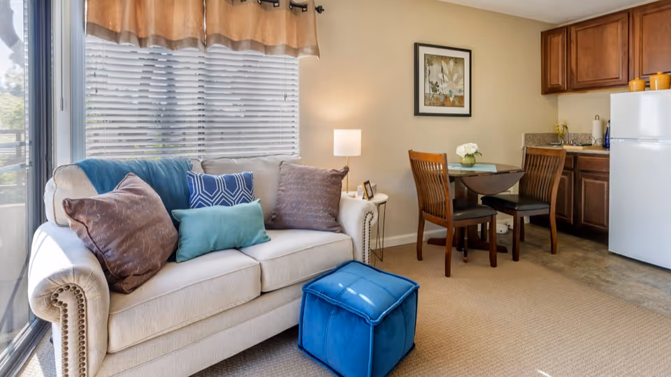 A cozy living area with a beige sofa adorned with multiple cushions in blue and brown tones, a blue ottoman in front, a small round dining table with two wooden chairs, a lamp on a side table, and a kitchenette with wooden cabinets and a white refrigerator.