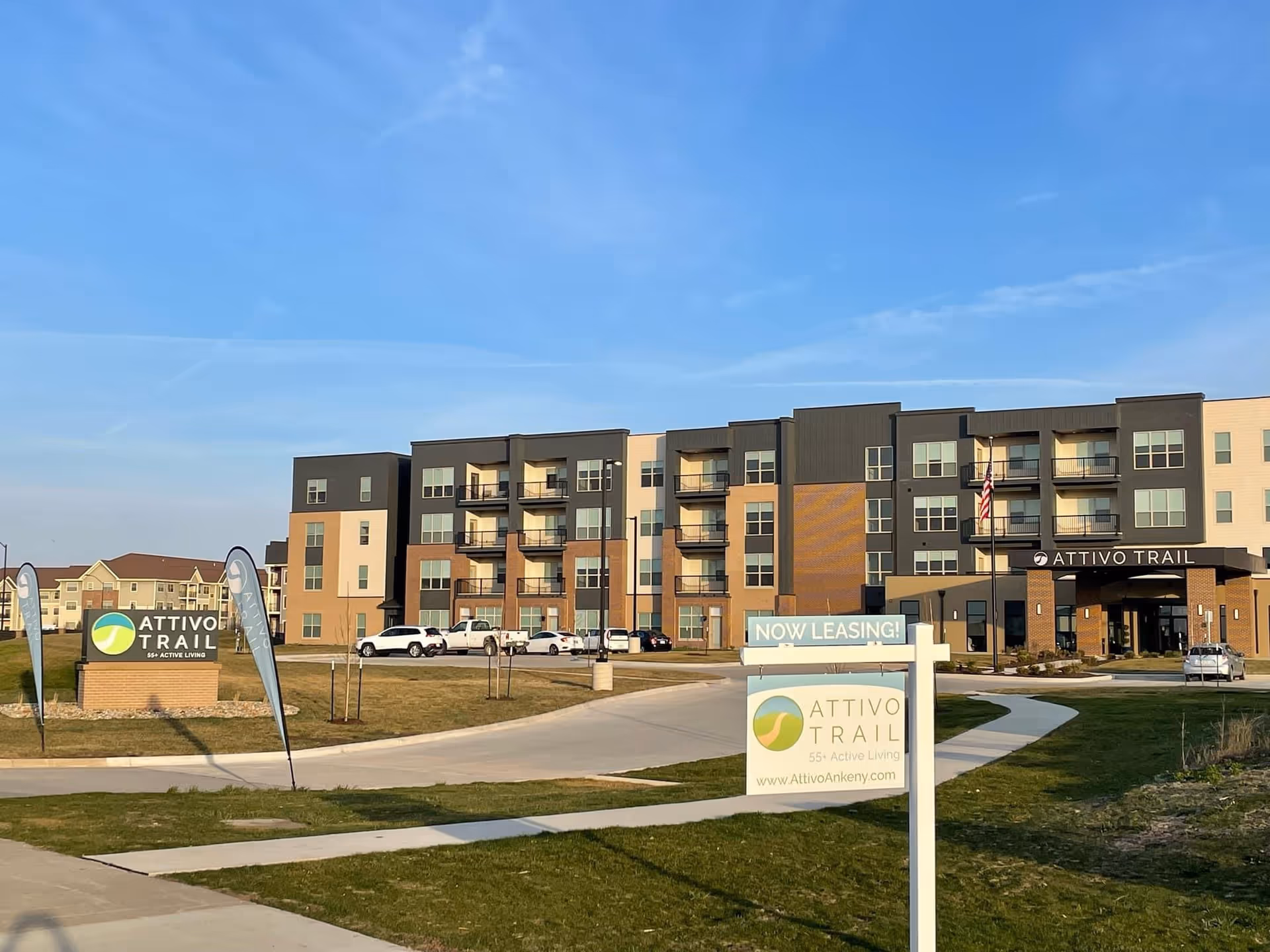 Front exterior of the Attivo Trail senior living building with entrance, signage, flags, and parked cars under a clear blue sky.