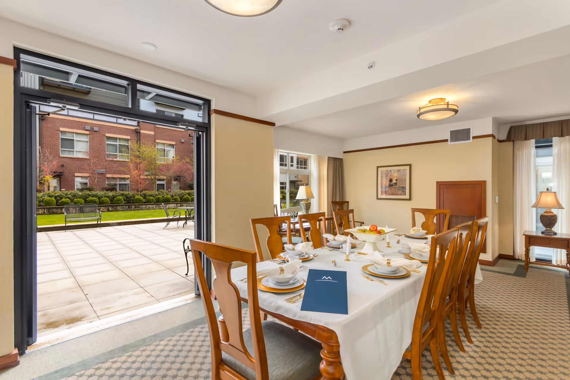 A dining room with a long wooden table set for a meal with white plates, napkins, and gold utensils. There are eight wooden chairs around the table. The room has beige walls, carpeted floor, and two lamps on side tables. Large glass doors open to an outdoor patio with benches and greenery, and a brick building is visible outside.
