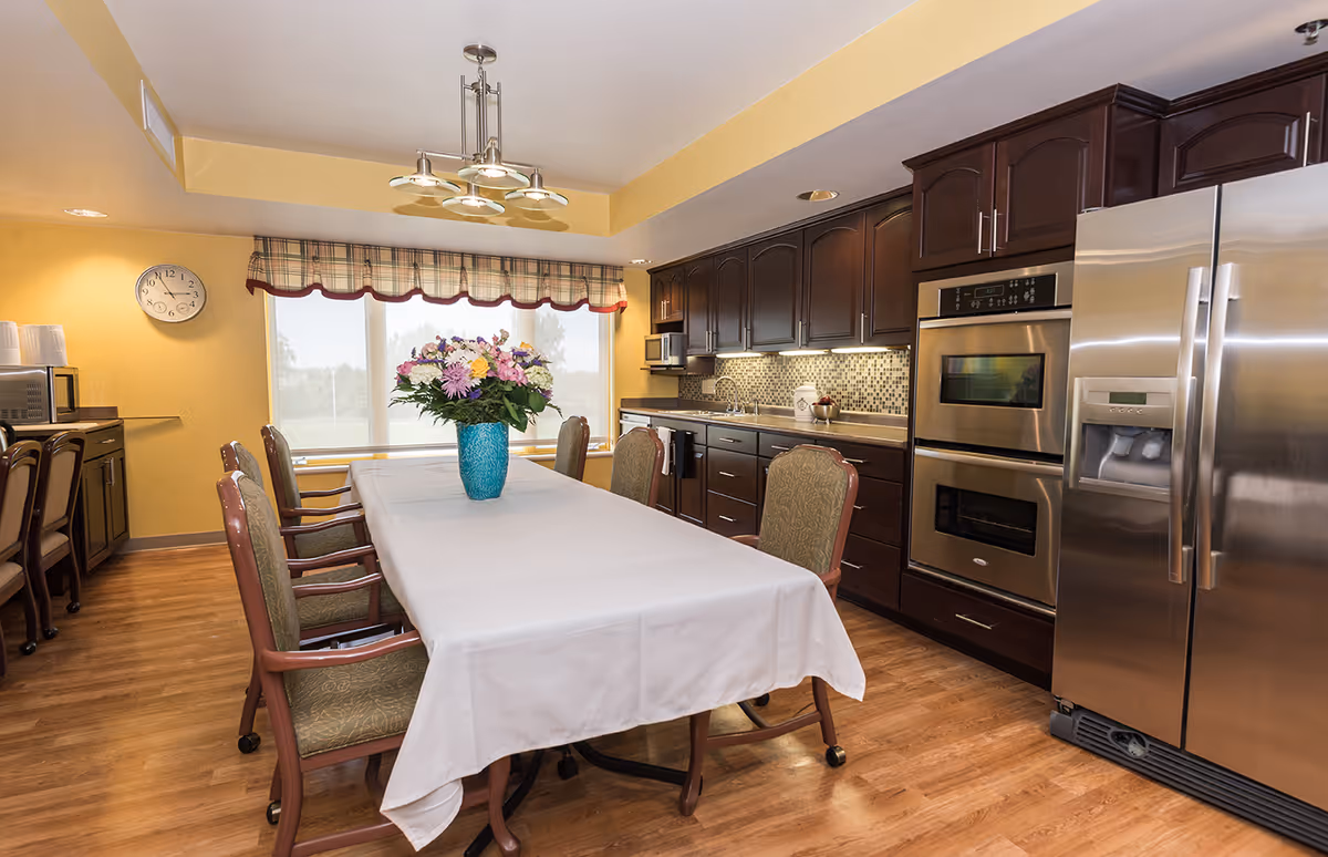A bright kitchen and dining area with a long table covered with a white tablecloth and surrounded by chairs. A vase with colorful flowers is placed in the center of the table. The kitchen features dark wooden cabinets, a stainless steel refrigerator, double ovens, a microwave, and a tiled backsplash. A clock is mounted on the yellow wall, and large windows with plaid valances allow natural light to fill the room.