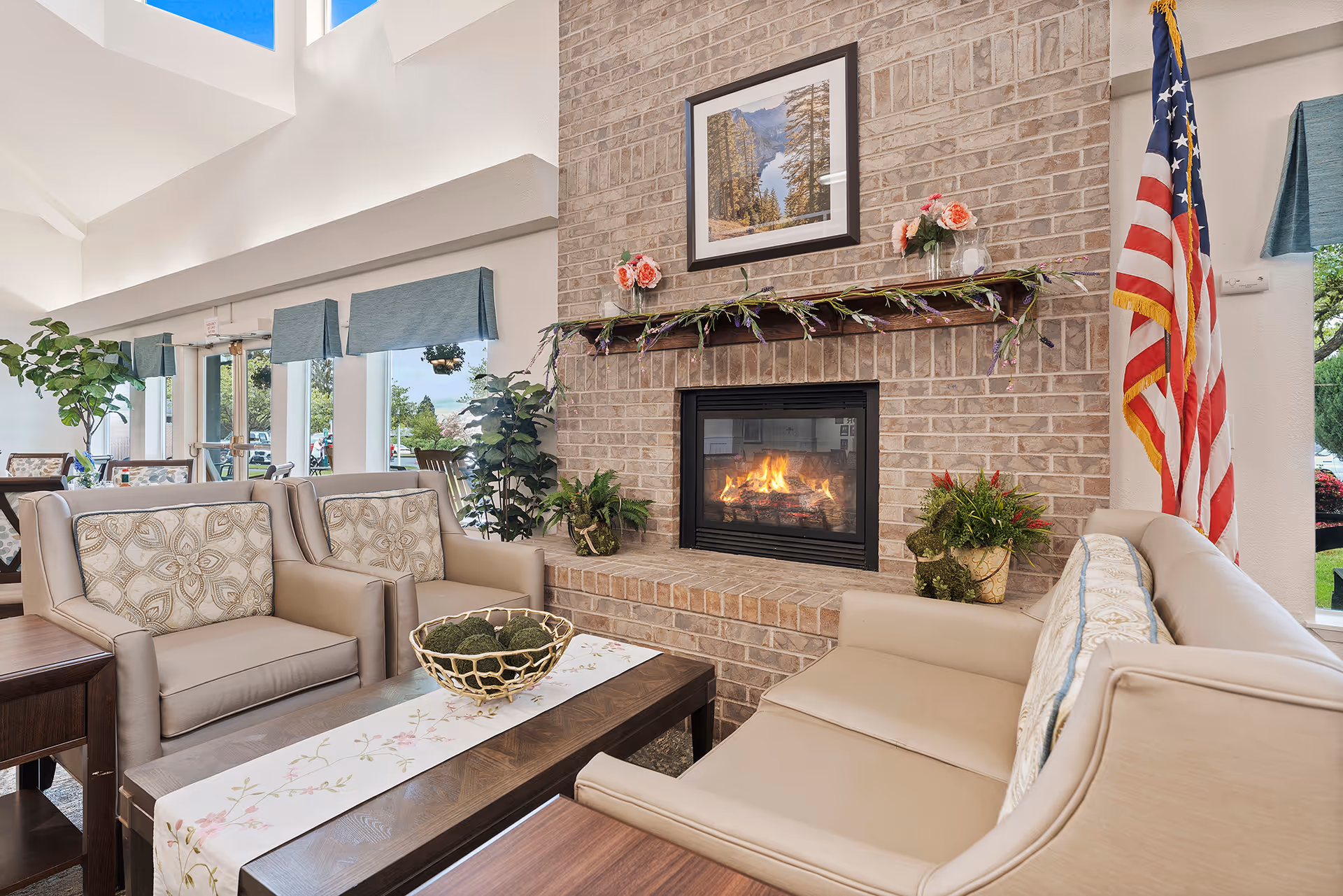 A cozy living room area with beige armchairs and a sofa arranged around a wooden coffee table with a decorative runner and a bowl of green moss balls. The room features a brick fireplace with a fire burning, decorated with flowers and a framed landscape photograph above it. There are large windows with blue valances letting in natural light, several potted plants, and an American flag displayed near the fireplace.