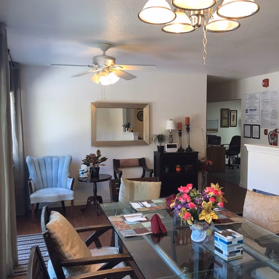 Glass-top dining table set with placemats and a floral centerpiece in a bright common room with chairs, a mirror, and side table.