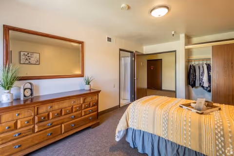 A bright bedroom with a wooden dresser topped by a large mirror, a bed with a yellow coverlet, an open closet, and a doorway to a bathroom.
