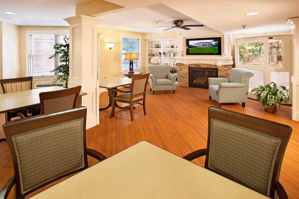 Sunlit senior living common room with tables and chairs in the foreground and armchairs around a fireplace with a wall-mounted TV in the background.