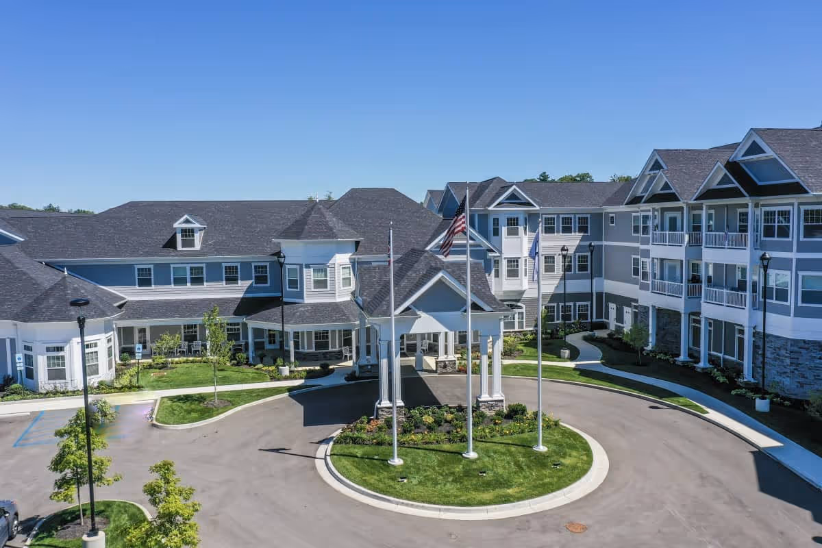 Front exterior of Traditions at Camargo showing the main entrance portico, flagpoles, circular driveway, and multi-story building under a clear blue sky.