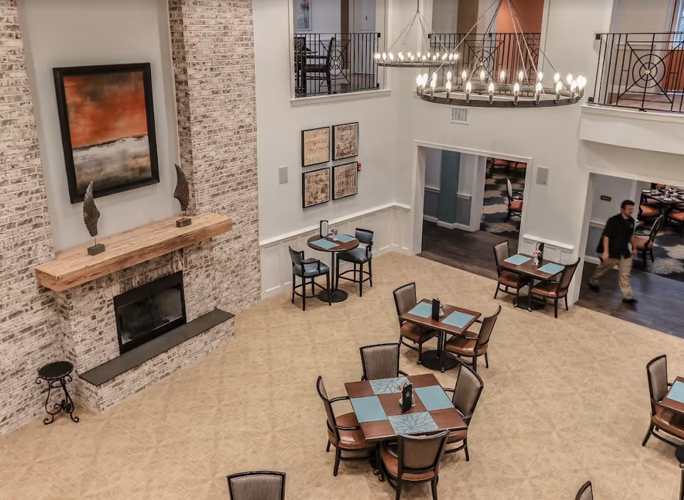 Interior view of a senior living facility dining area with several tables and chairs arranged on a beige carpet. There is a large brick fireplace with a wooden mantel decorated with two sculptures and a framed abstract painting above it. The room has high ceilings with two large circular chandeliers and a balcony railing visible on the upper level. A person is walking through a doorway leading to another dining area with more tables and chairs.
