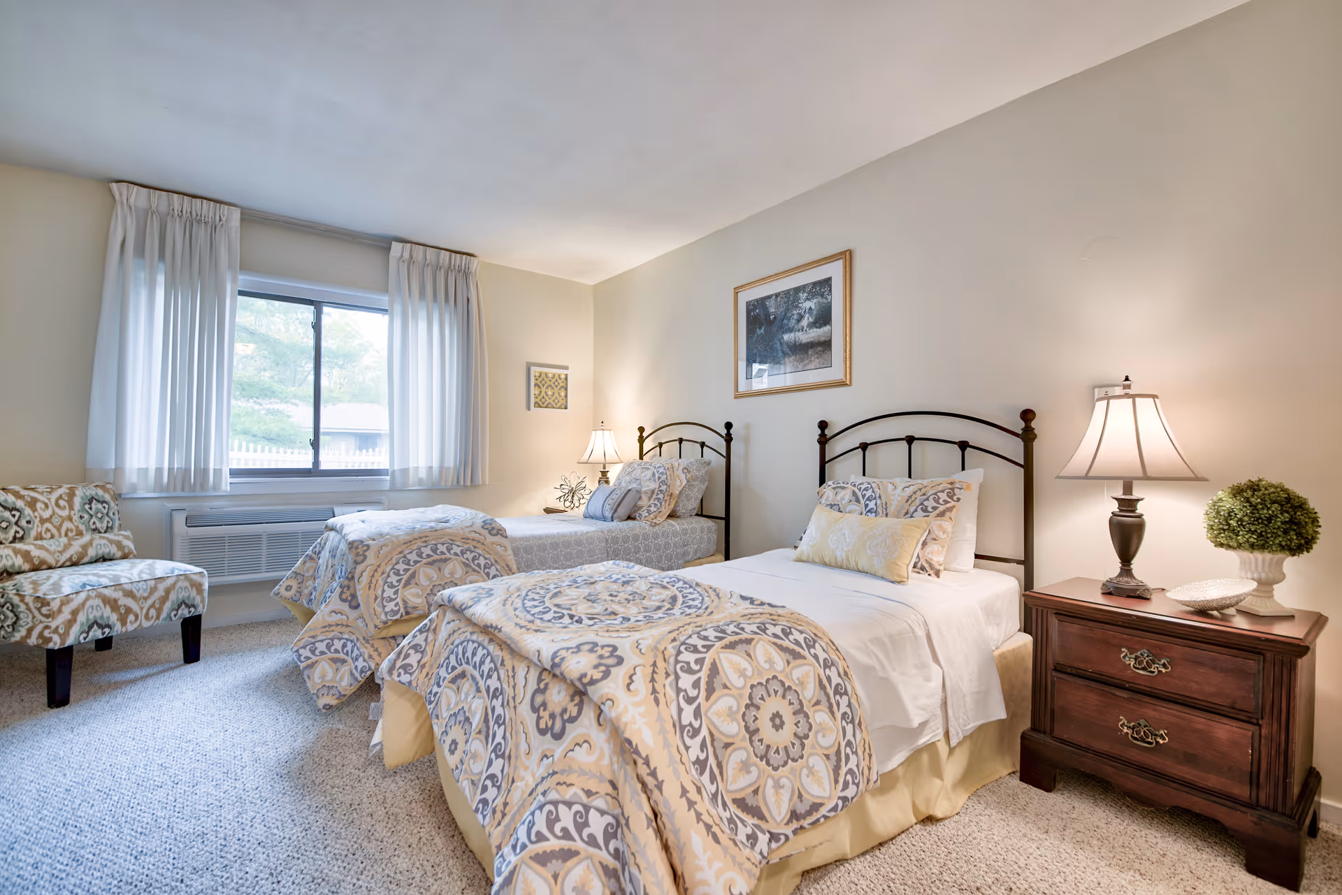 A bright and cozy bedroom with two twin beds featuring patterned bedspreads in beige, blue, and white tones. Each bed has a black metal headboard and decorative pillows. Between the beds is a wooden nightstand with a lamp, a small potted plant, and a decorative dish. A window with white curtains allows natural light into the room. There is also an upholstered chair with a patterned fabric near the window. The walls are light-colored with framed artwork hanging above the beds.