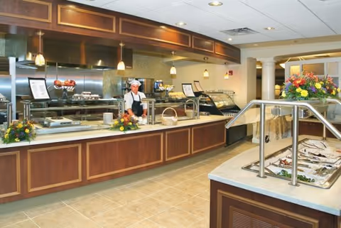 A clean and well-lit cafeteria or dining area with a long wooden serving counter. A staff member wearing a white chef hat and apron stands behind the counter. There are food display cases, a salad bar, and decorative flower arrangements on the counters. The ceiling has recessed lighting and pendant lights above the serving area.