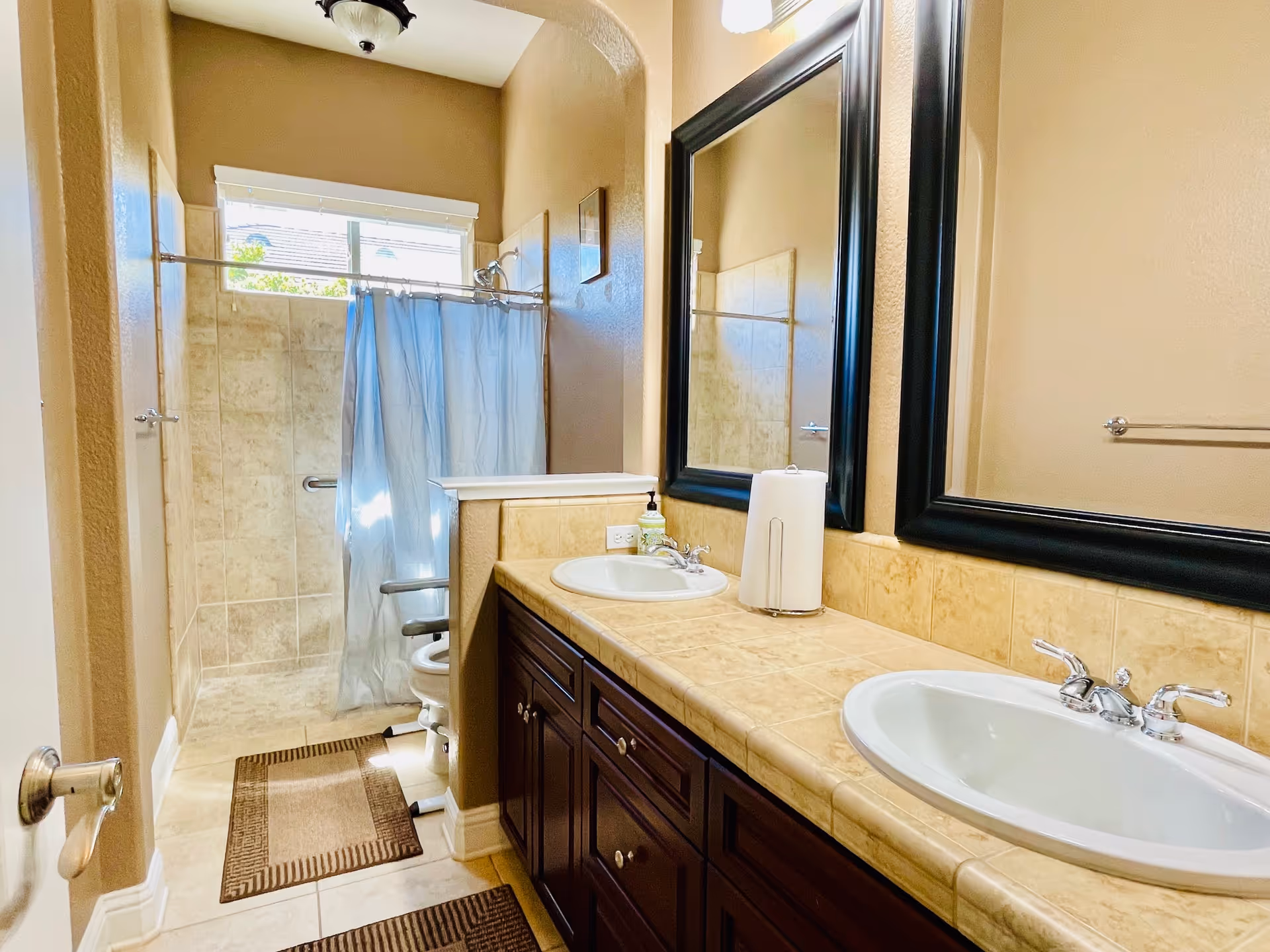 A bathroom with two sinks set in a tiled countertop with dark wood cabinets below. Above the sinks are two large framed mirrors. To the left is a walk-in shower with beige tiles, a silver grab bar, and a blue shower curtain. The bathroom has beige walls and two brown floor mats on the tiled floor.