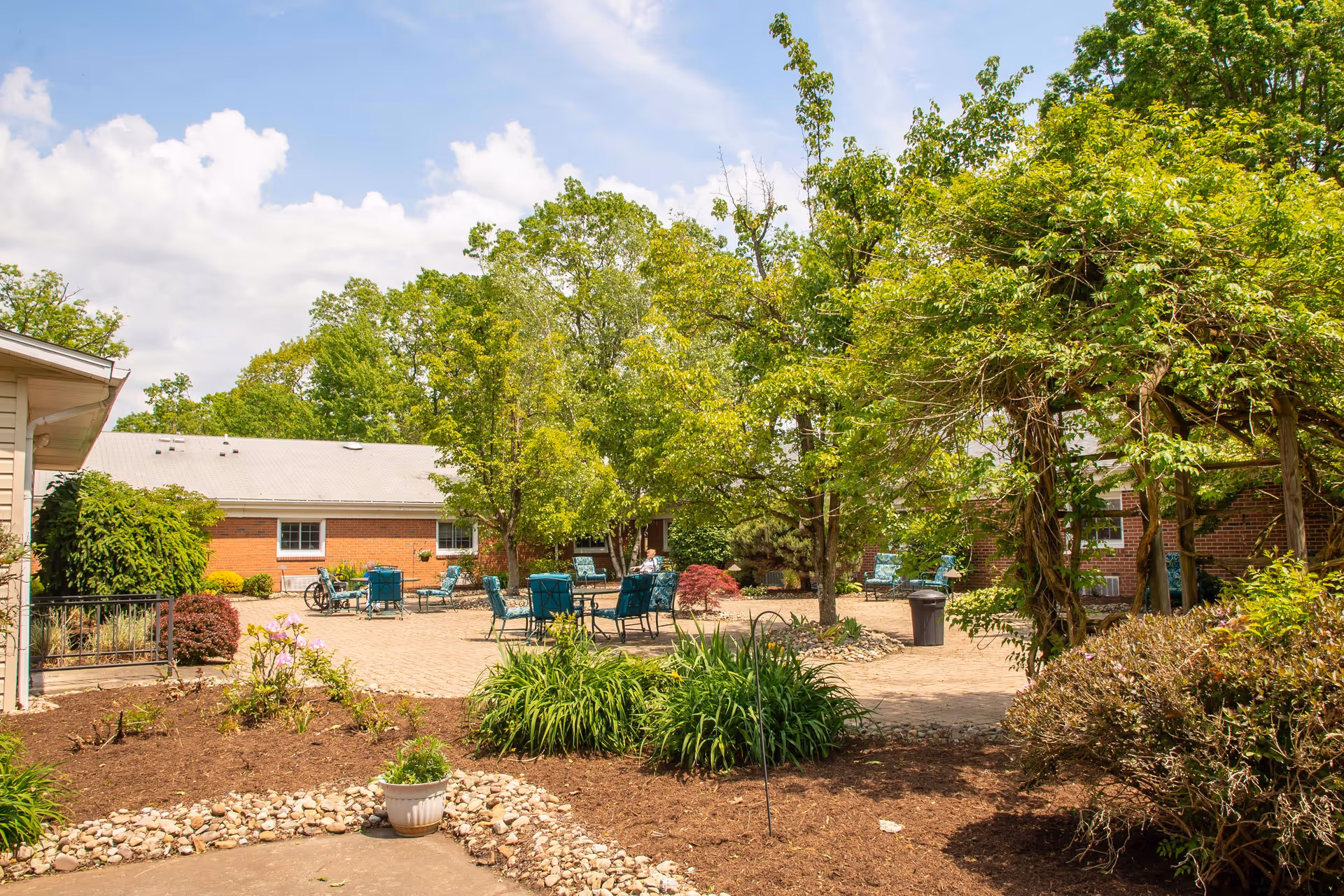 Outdoor courtyard area with several green trees, shrubs, and plants. There are multiple blue metal chairs and tables arranged on a paved surface. A brick building with white-framed windows is visible in the background under a partly cloudy sky.