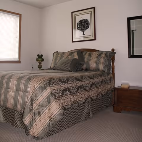 A neatly made bed with green and beige patterned bedding, wooden headboard, nightstands, a lamp, framed art, and a window in a simple bedroom.