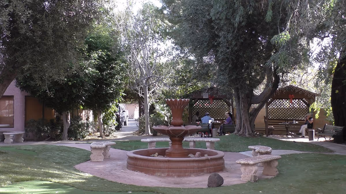 A peaceful outdoor garden area at Ambassador Garden Retirement featuring a central tiered water fountain surrounded by stone benches. There are large trees providing shade and two gazebo-like structures with lattice walls where people are seated and conversing.