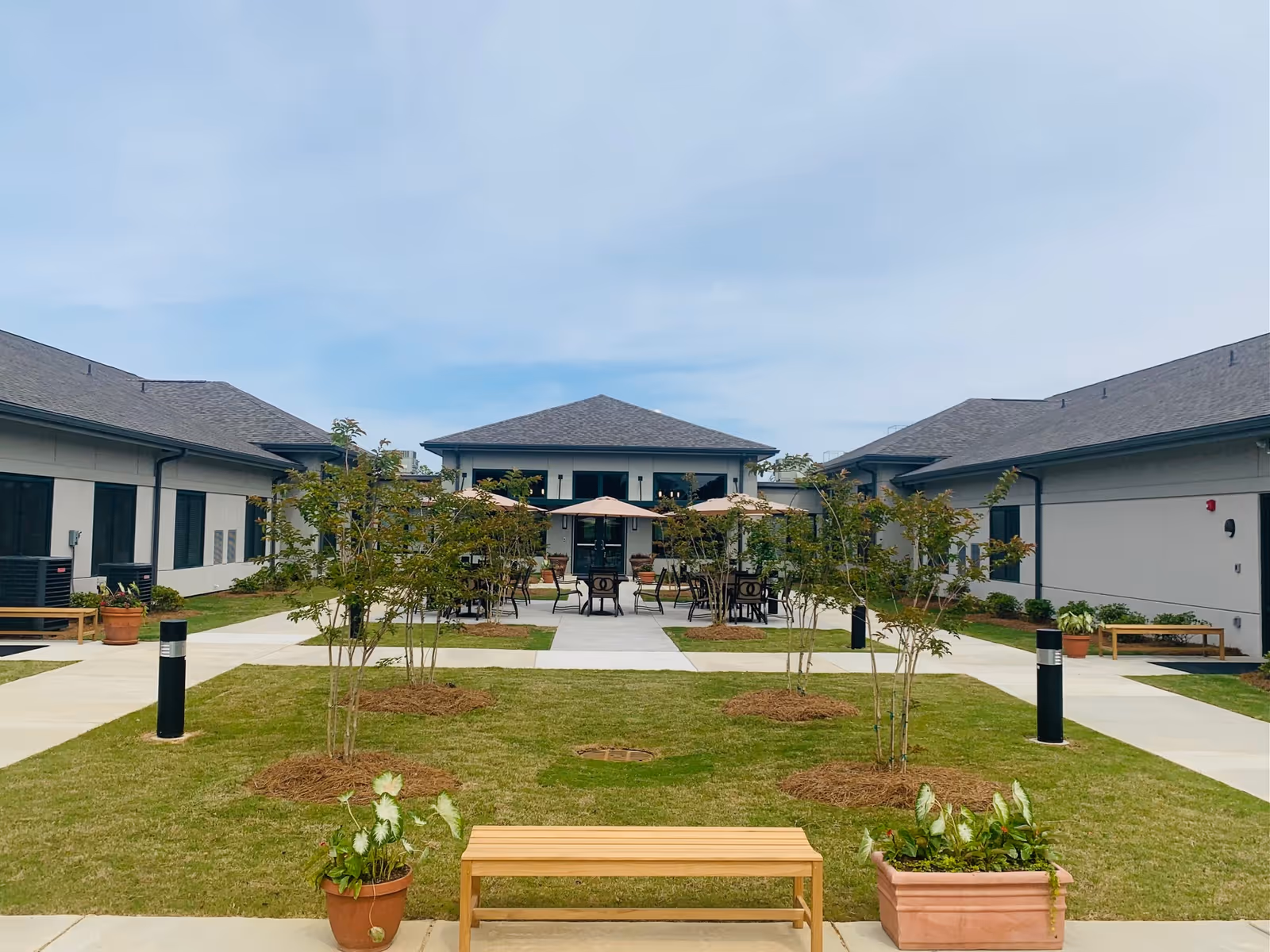Outdoor courtyard area of a senior living facility with green grass, small trees, potted plants, wooden benches, and tables with umbrellas. The courtyard is surrounded by single-story buildings with gray roofs under a partly cloudy sky.