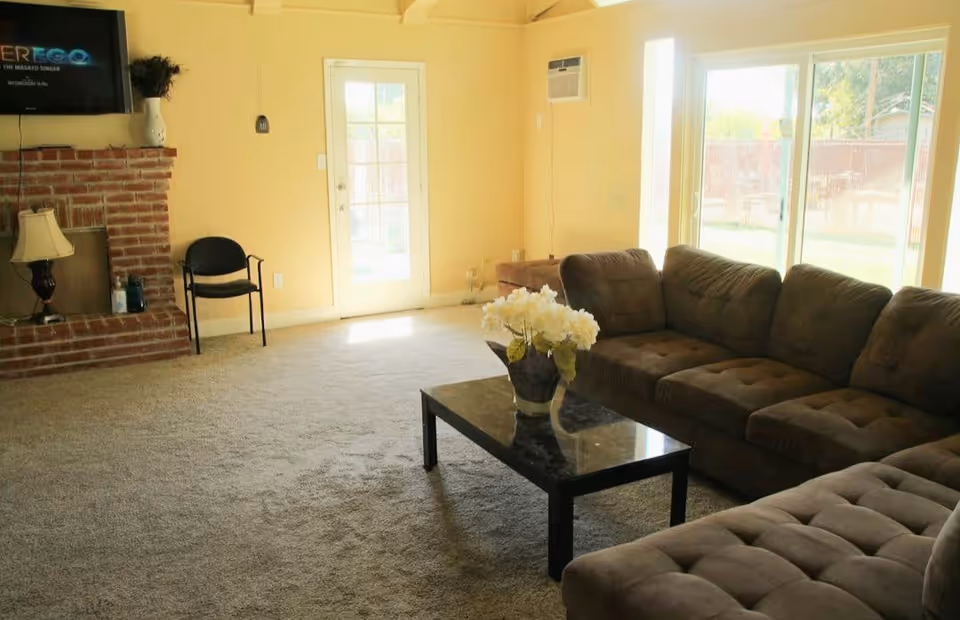 A cozy living room with a large brown sectional sofa, a black coffee table with a vase of white flowers, a brick fireplace with a lamp and decorative items, a black chair, a wall-mounted TV, a door with glass panels, and large windows letting in natural light.