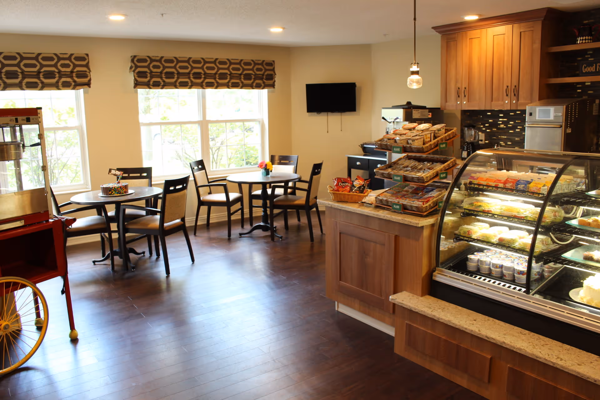 A cozy dining area with several round tables and chairs near large windows with patterned valances. To the right, there is a wooden counter displaying various snacks and a glass refrigerated display case filled with packaged food items. The room has warm lighting and wooden flooring.