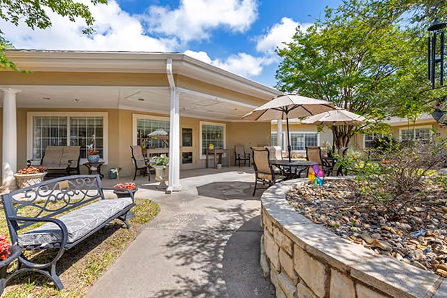 Outdoor courtyard patio with tables, chairs and umbrellas beside a covered walkway and stone planter in front of a single-story building.