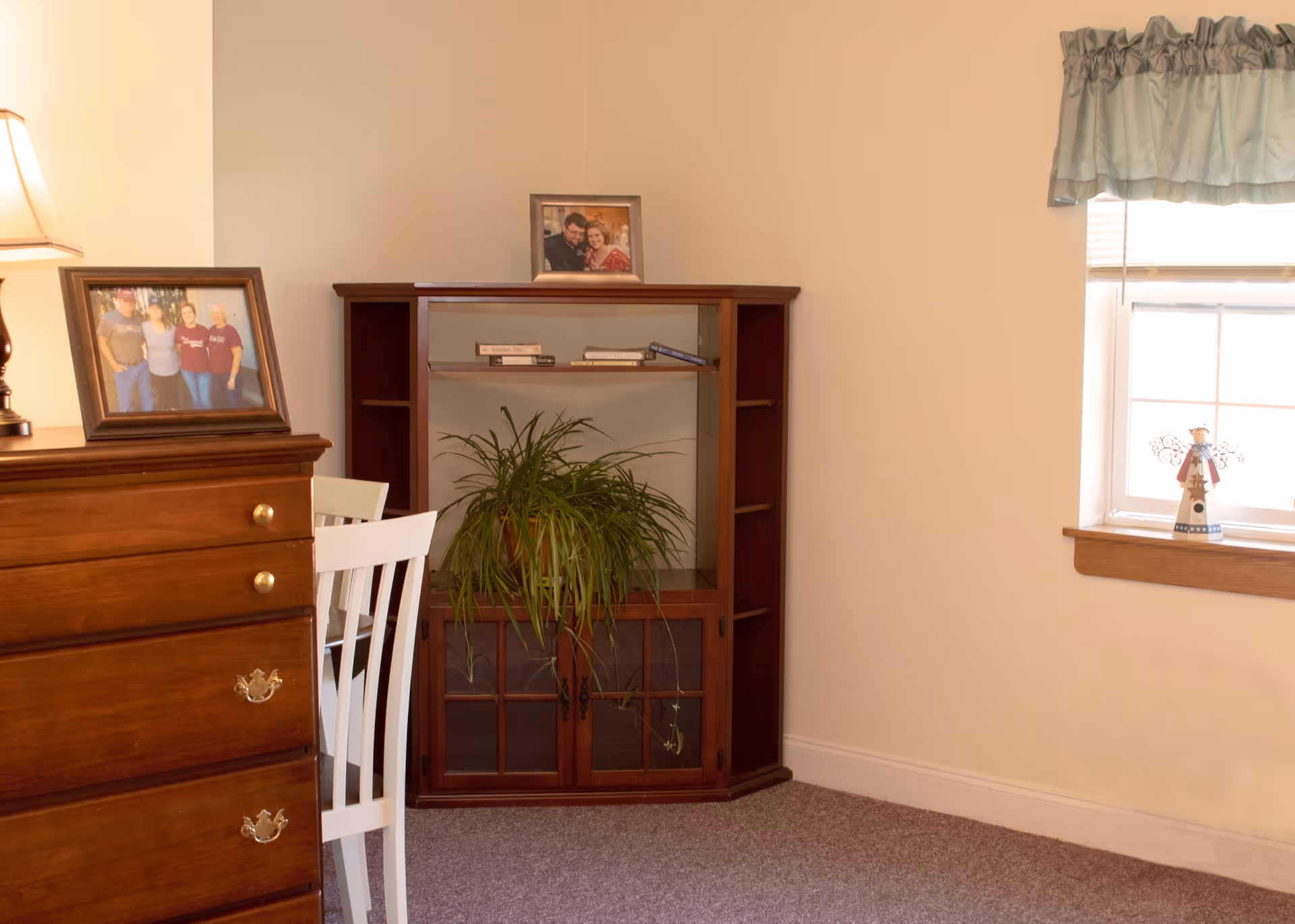 A corner of a room with a wooden dresser on the left holding a framed photo and a lamp. Next to it is a white chair and a wooden cabinet with glass doors containing a green potted plant and some books on the top shelf. A window with a light blue valance is on the right side with a small angel figurine on the windowsill.