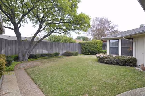 Well-kept backyard with green lawn, trees, shrubs, a fence, and house exterior.