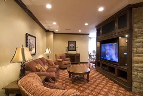 A cozy living room in a senior living community featuring a patterned carpet, a round wooden coffee table, a sofa and armchairs with cushions, a wooden entertainment center with a television, table lamps, framed artwork on the walls, and recessed ceiling lights.