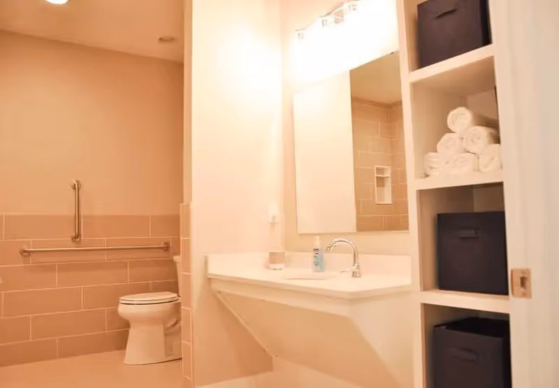 A clean and well-lit bathroom featuring a toilet with grab bars on the wall, a white sink with a faucet, a large mirror above the sink, and built-in shelving holding rolled white towels and dark storage bins.