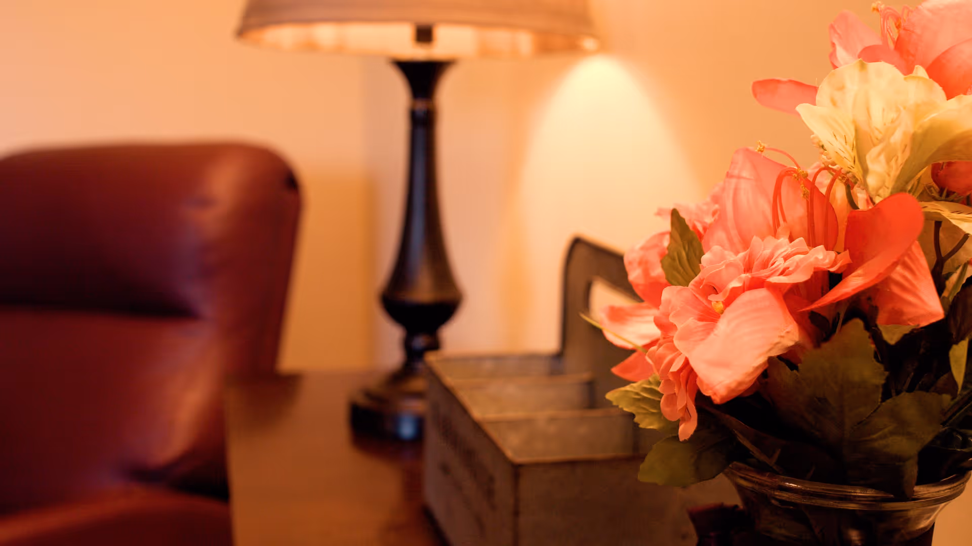 Close-up view of a table with a decorative vase of pink and white flowers, a black table lamp with a beige shade, and a blurred brown leather chair in the background.
