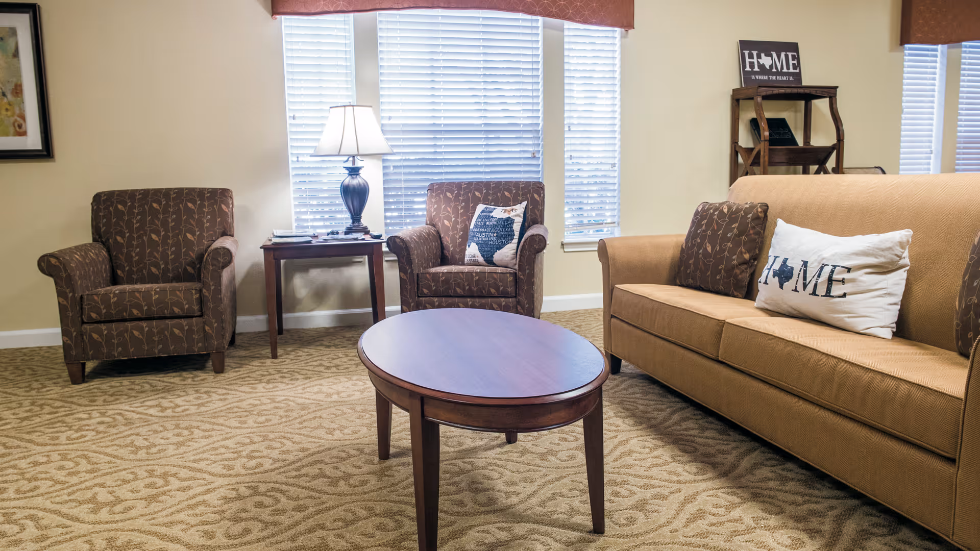 A cozy living room with a beige sofa and two patterned armchairs. The sofa has decorative pillows, one with the word 'HOME' and a Texas state silhouette. A wooden oval coffee table is in the center on a patterned carpet. Behind the armchairs is a wooden side table with a lamp, and windows with white blinds letting in natural light. A small wooden shelf with a decorative sign is in the corner.