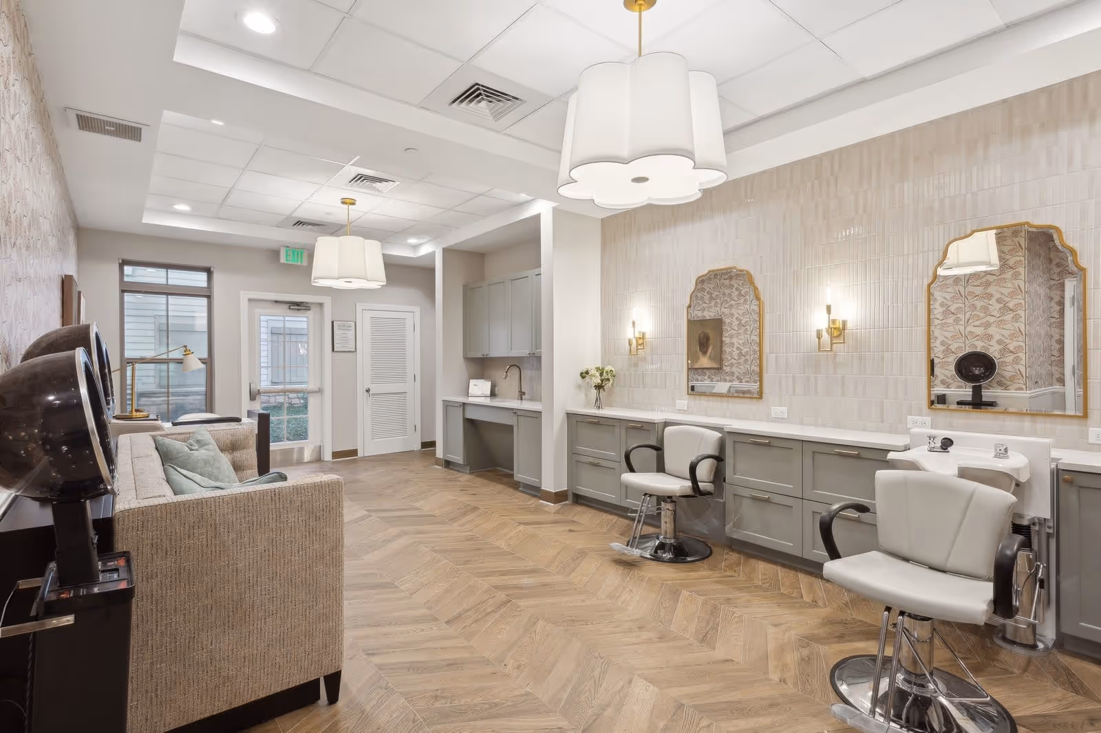 Interior view of a senior living facility salon area with two salon chairs in front of mirrors mounted on a tiled wall. There is a beige couch with pillows on the left side, two hair dryers, and cabinetry with a sink. The room has wood-patterned flooring, pendant lighting, and a door leading outside.