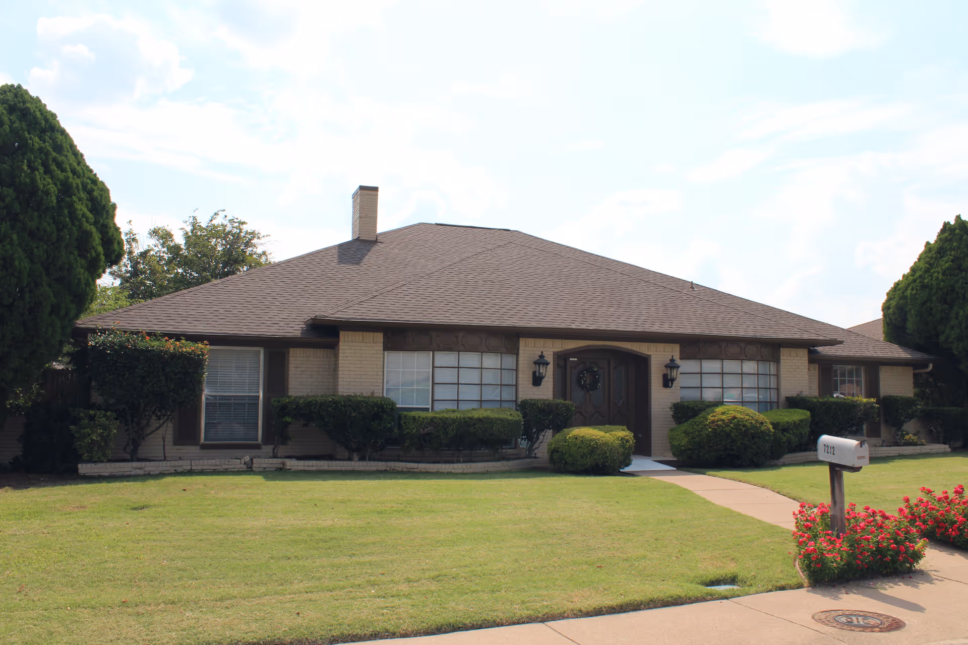 Single-story brick house with a brown shingled roof, front door with a wreath, multiple windows, neatly trimmed bushes, green lawn, a sidewalk leading to the entrance, and a mailbox with red flowers nearby under a partly cloudy sky.