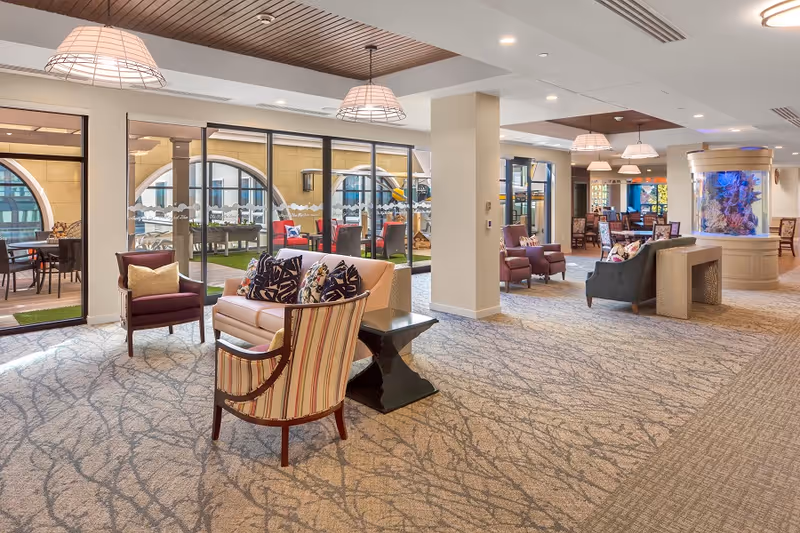 A spacious and well-lit senior living common area with comfortable seating including armchairs and sofas adorned with decorative pillows. Large windows and glass doors reveal an outdoor patio with additional seating. The room features a large cylindrical aquarium filled with colorful fish, modern pendant lighting, and a patterned carpeted floor.