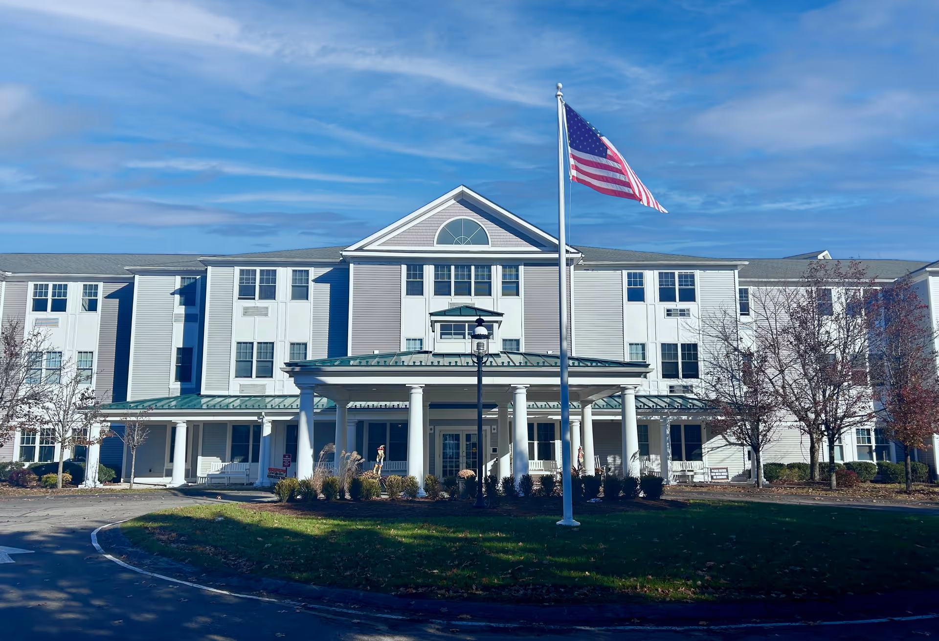 Front exterior view of a large, three-story senior living facility building with white and light gray siding, a covered entrance with columns, an American flag on a flagpole in front, and a circular driveway surrounded by grass and trees under a blue sky.