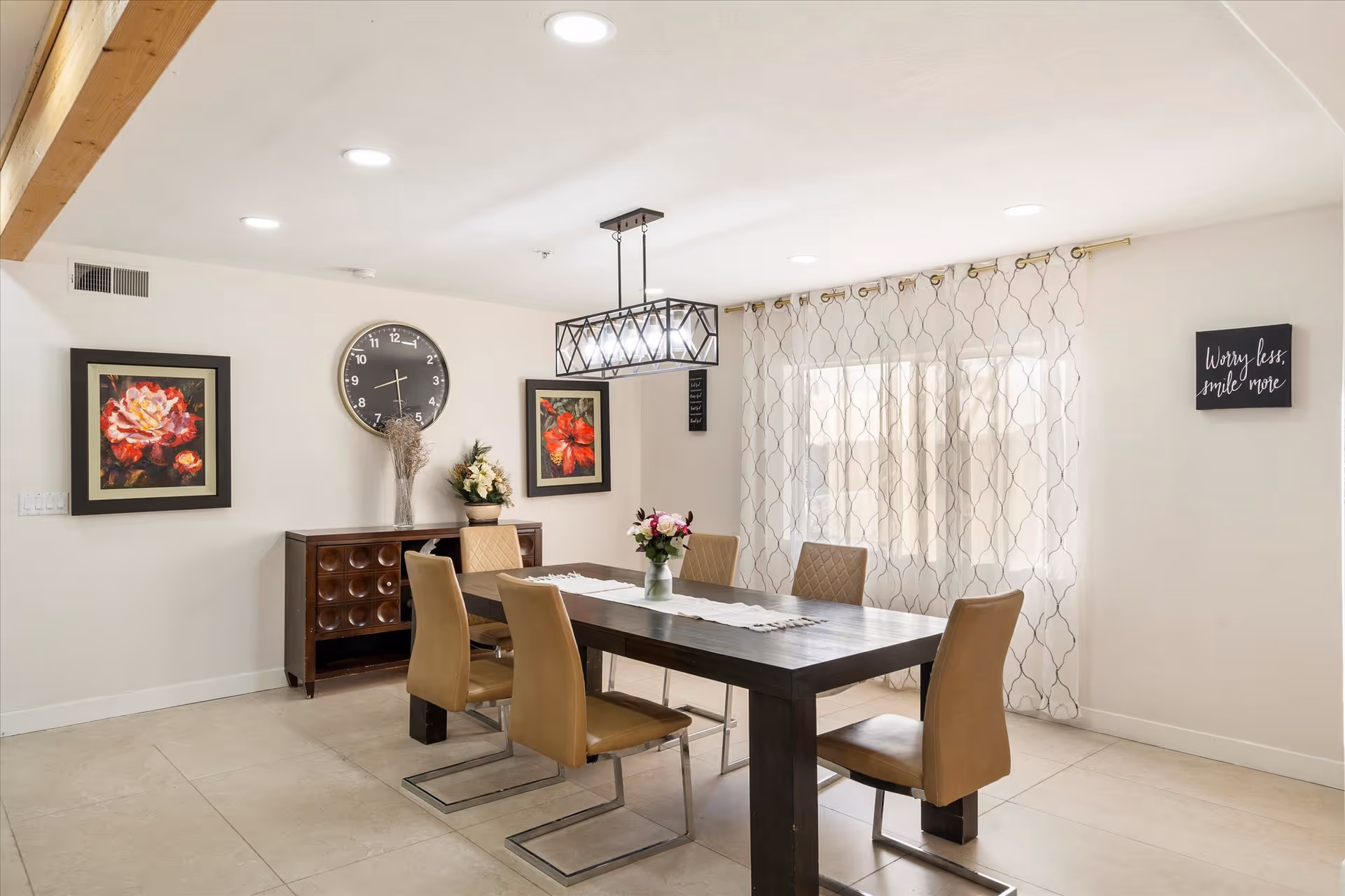A bright dining room with a dark wooden table surrounded by six tan chairs. The room features a modern rectangular chandelier above the table, two framed floral paintings on the wall, a large round clock, and a sideboard with decorative flowers. Sheer patterned curtains cover a window, and a small wall sign reads 'Worry less, smile more.'