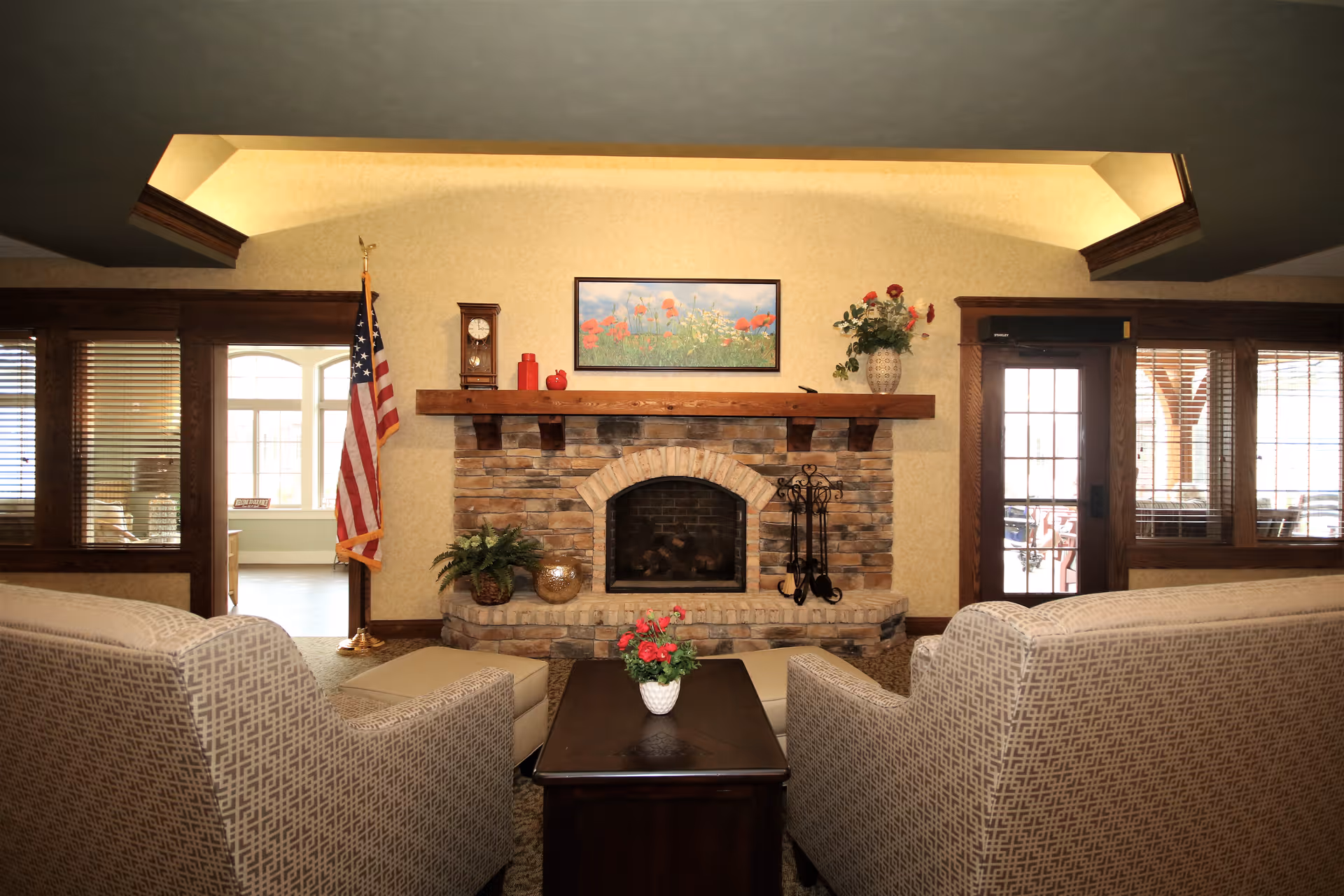 Cozy living room with two patterned armchairs facing a stone fireplace. The fireplace mantel is decorated with a clock, red decorative items, and a vase with flowers. An American flag stands to the left of the fireplace. There is a framed picture of flowers hanging above the mantel. The room has wooden trim around windows and doors, with blinds on the windows and a glass door on the right side.