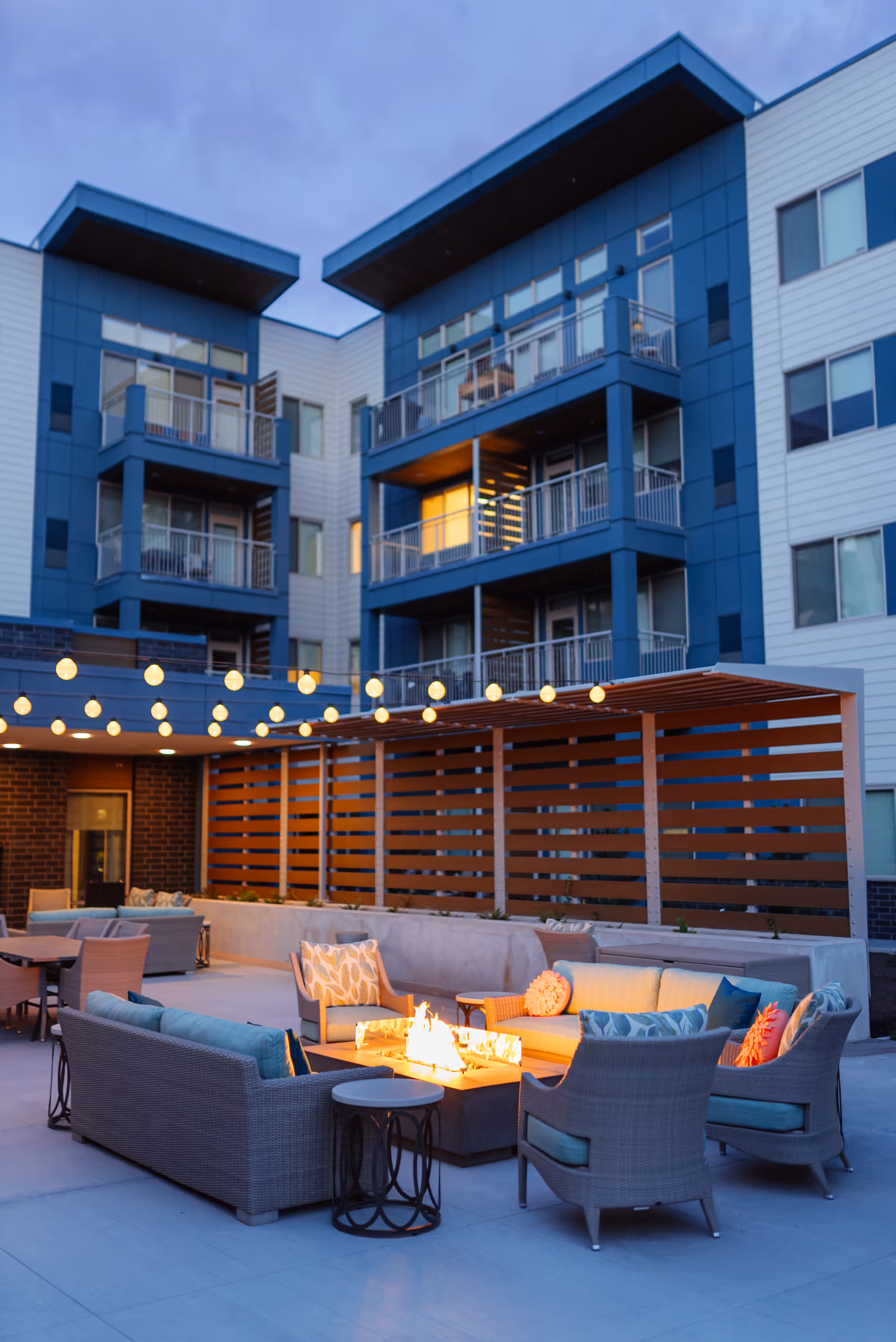 Outdoor patio area at dusk with modern wicker seating arranged around a rectangular fire pit. String lights hang overhead, and a multi-story building with balconies is visible in the background.