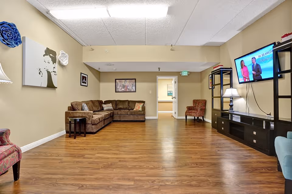 Spacious common living room with a sectional sofa, armchairs, wall-mounted TV and shelving over wood floors.