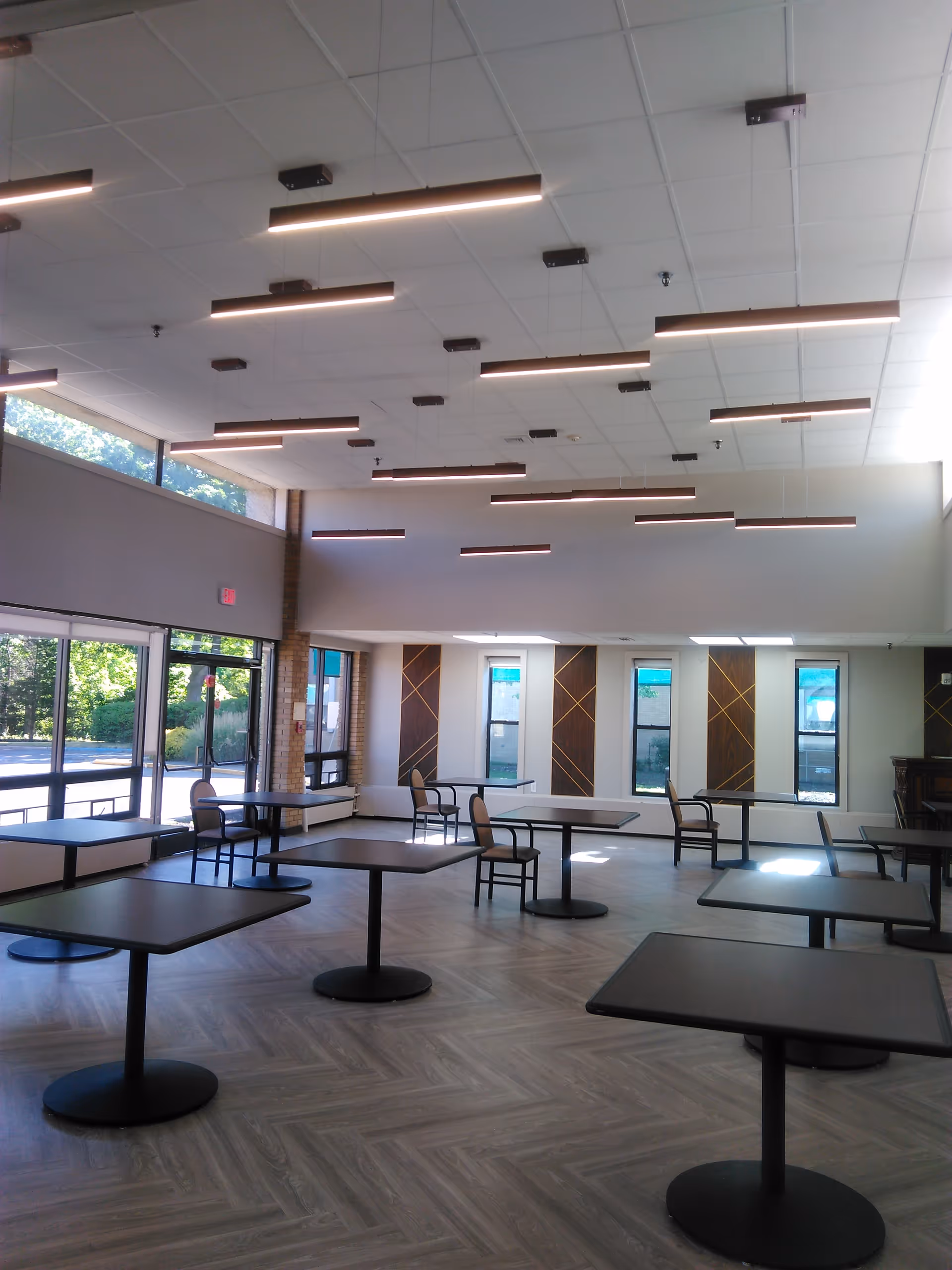 A spacious dining area with multiple square tables and chairs arranged neatly. The room has large windows allowing natural light to enter, modern ceiling lights, and a patterned wooden floor. The walls feature decorative panels between the windows.