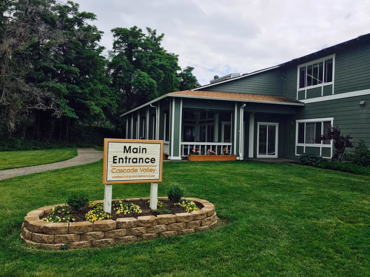 Exterior view of a green two-story building with white trim, surrounded by a well-maintained lawn and trees. In front of the building is a sign that reads 'Main Entrance Cascade Valley Assisted Living and Memory Care' placed in a small flower bed with yellow flowers.