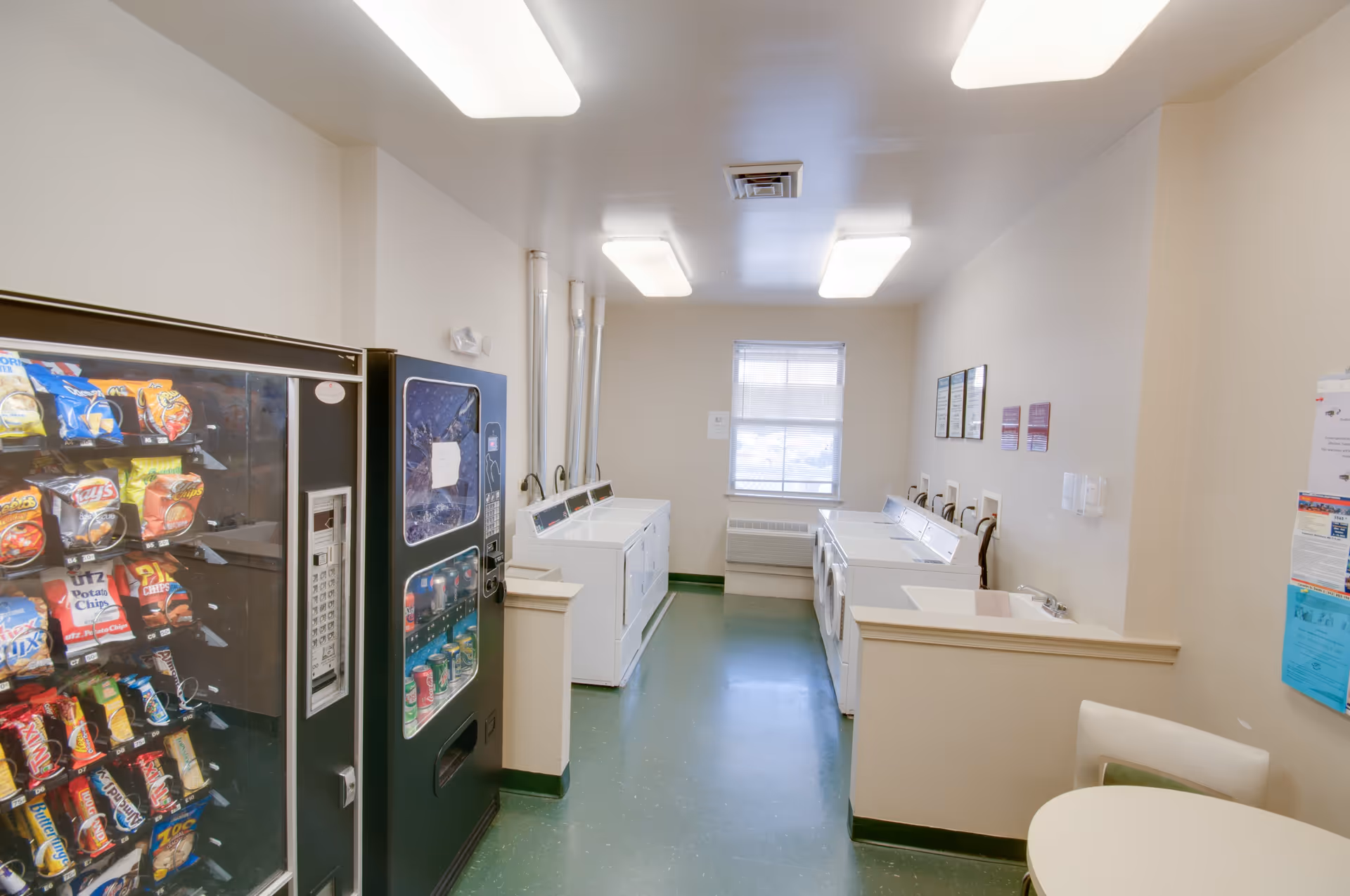 A laundry room with several white washing machines and dryers along the walls. There is a vending machine with snacks and another vending machine with drinks on the left side. The room has a green floor, beige walls, and fluorescent ceiling lights. A small round table with chairs is partially visible on the right side.