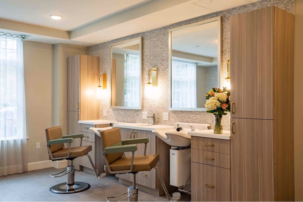 A well-lit salon area with two brown and green salon chairs in front of a counter with two sinks and large mirrors. The counter has light wood cabinetry, wall-mounted lights, and a vase with a bouquet of flowers. Sheer curtains cover the windows, allowing natural light to fill the room.