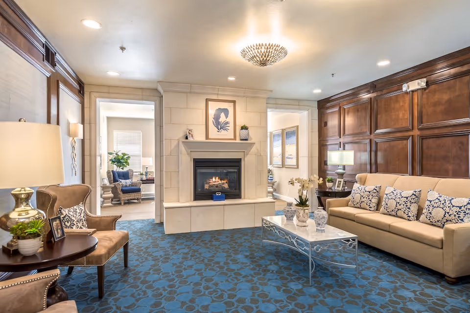 A cozy senior living facility lounge area with a beige sofa adorned with patterned cushions, two armchairs, a marble coffee table with decorative vases, and a fireplace with a framed artwork above it. The room features wood-paneled walls, a blue patterned carpet, and warm lighting from lamps and a ceiling fixture.