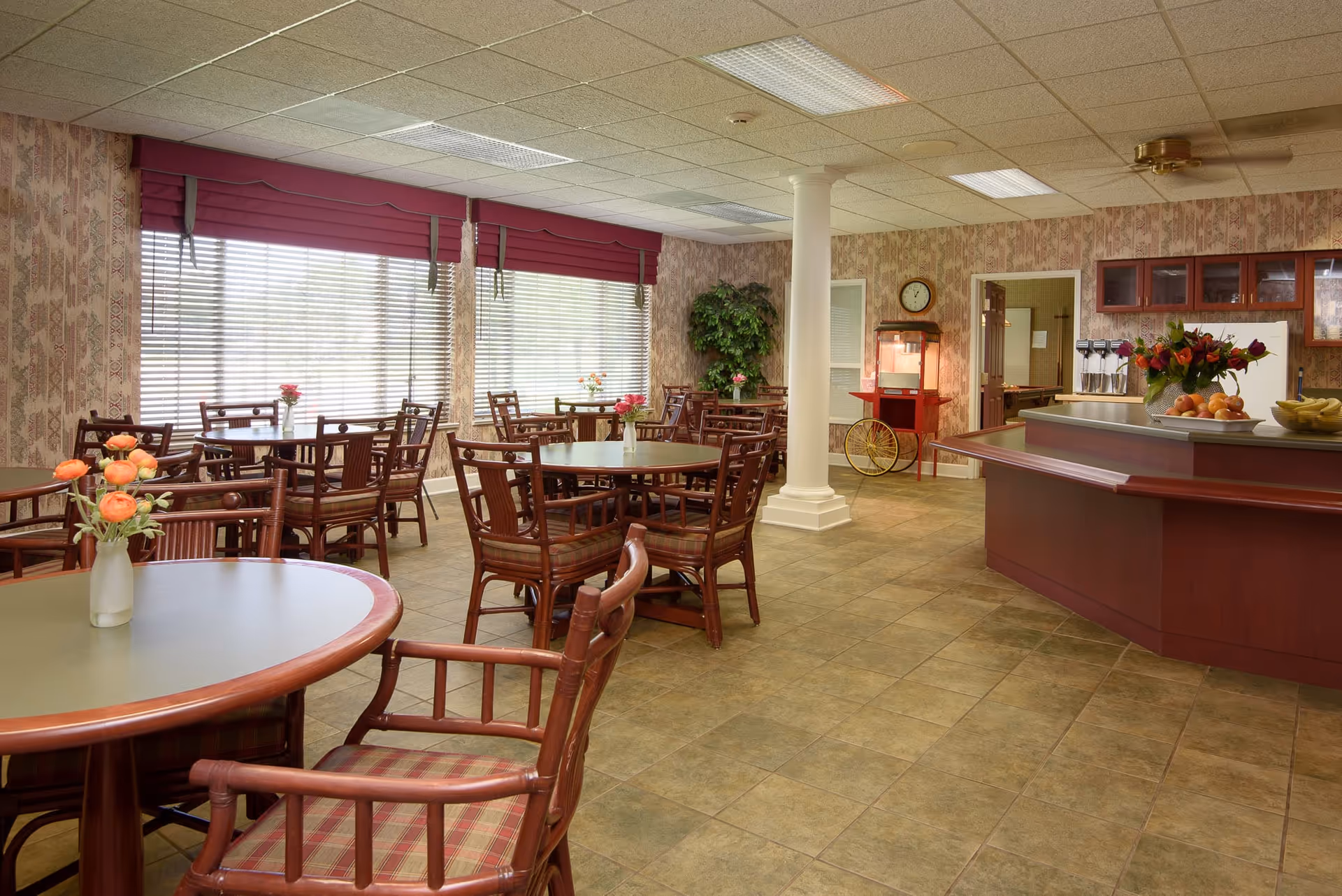 A dining room with several round tables and wooden chairs with plaid cushions. Each table has a small vase with flowers. Large windows with blinds and red valances let in natural light. There is a white column in the middle of the room, a popcorn machine on a red cart, and a counter with fruit bowls and a flower arrangement. The walls have patterned wallpaper and the floor is tiled.