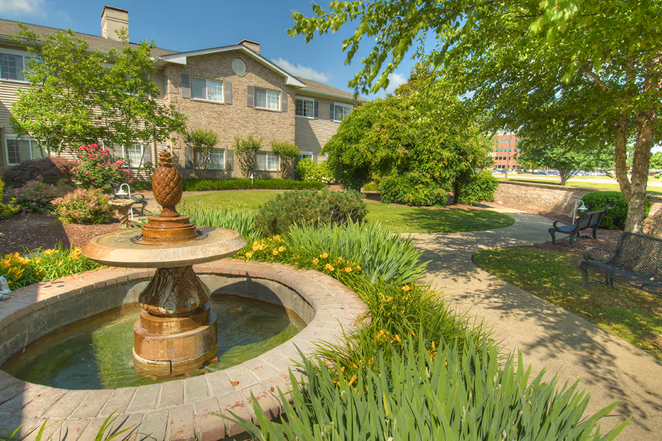 A landscaped courtyard with a decorative fountain, paved walkway, benches, and a two-story building in the background.