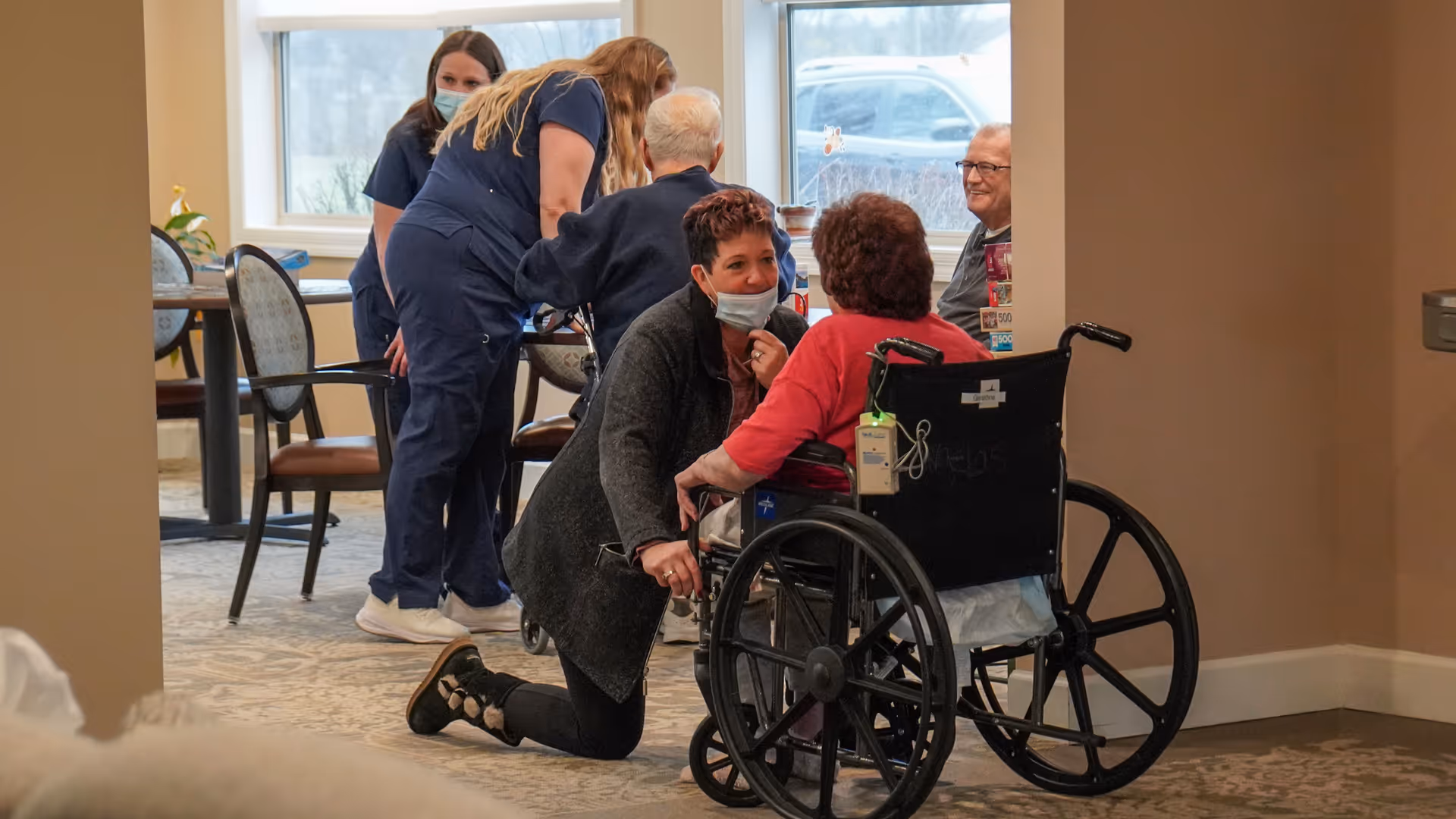 A caregiver kneels to speak with a resident in a wheelchair in a communal dining area while staff and other residents sit and stand nearby.