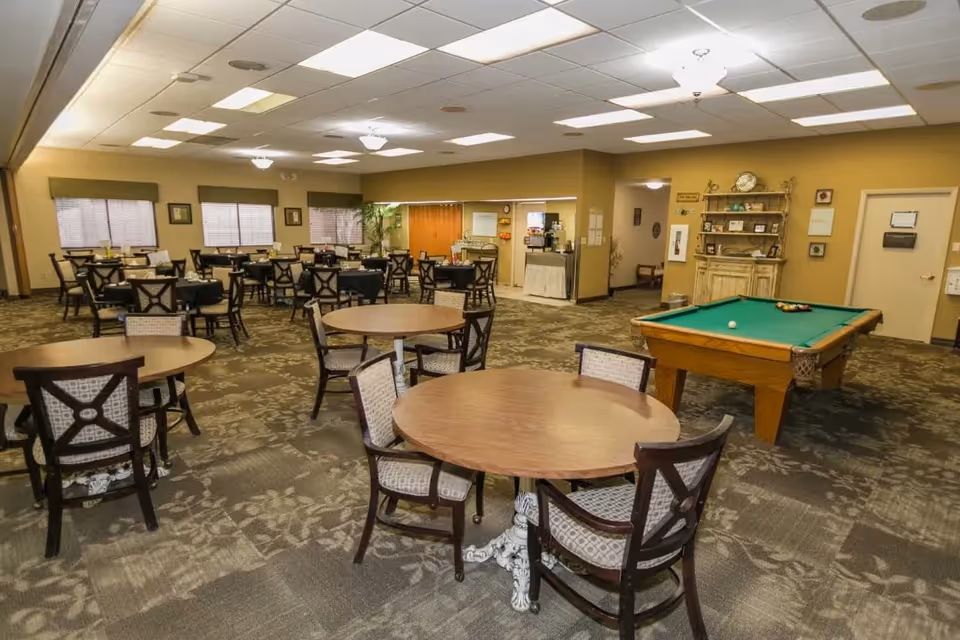 A spacious senior living facility common area with multiple round wooden tables and chairs arranged for seating. There is a pool table with billiard balls set up on the right side of the room. The room has beige walls, patterned carpet, and several windows with blinds. A wooden cabinet with decorative items is visible against the far wall, and there is a coffee station in the background.
