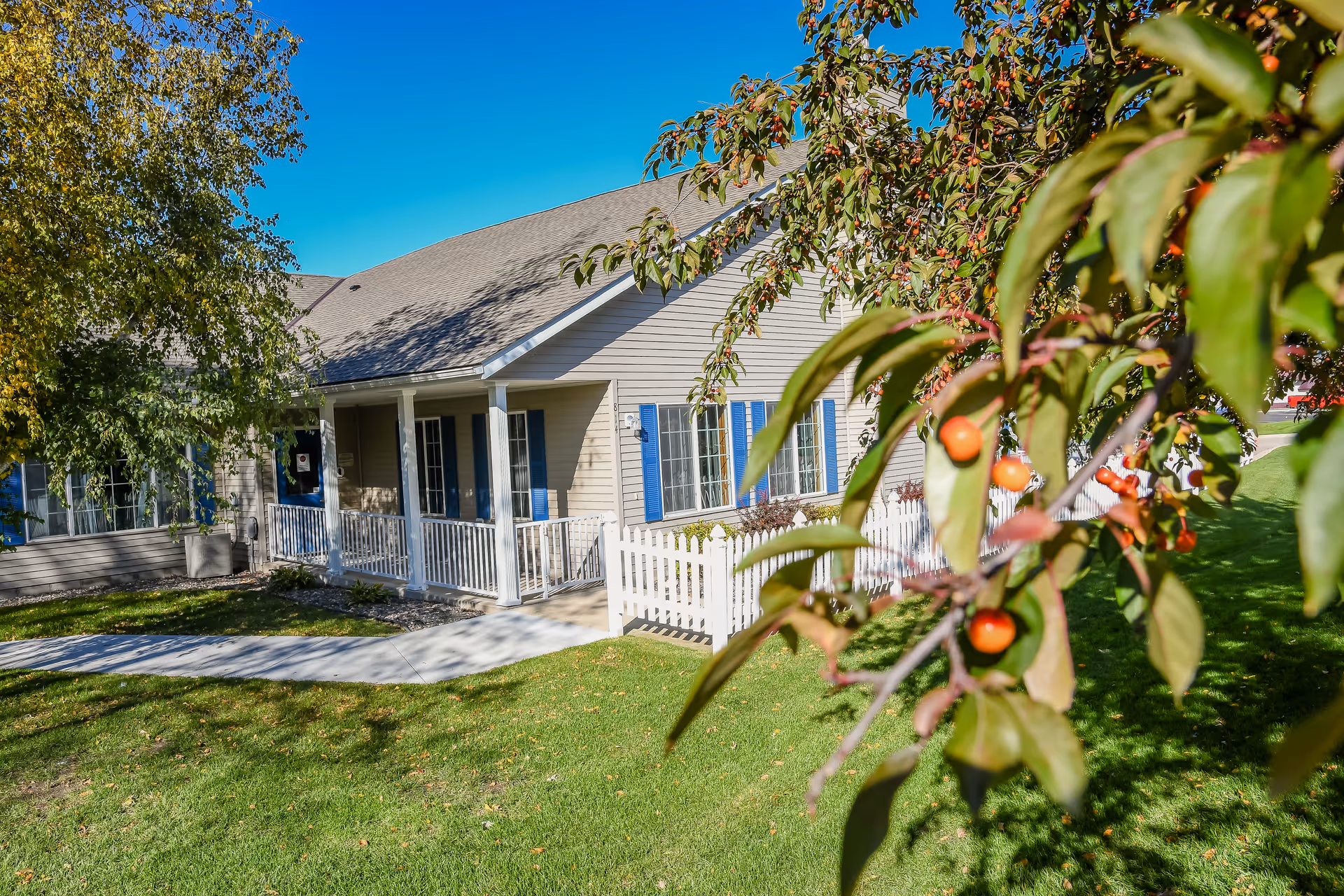 A single-story residential building with beige siding and blue shutters, featuring a white picket fence and a covered porch with white railings. The building is surrounded by green grass and trees, with a clear blue sky overhead. In the foreground, branches with orange berries are visible.