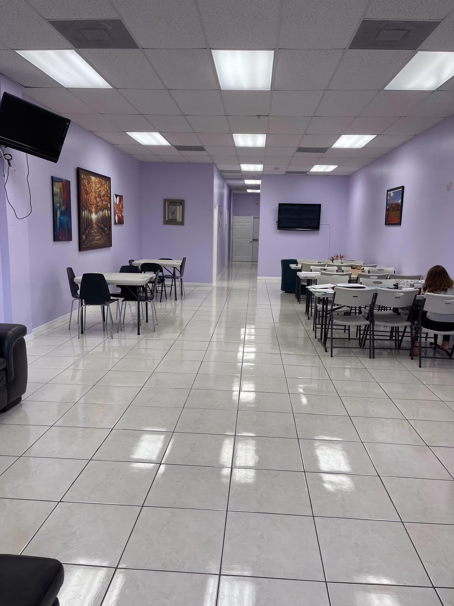 Interior view of a spacious room with light purple walls and white tiled floor. The room contains several tables and chairs arranged on both sides, with a few paintings on the walls and two wall-mounted televisions. A person is seated at one of the tables on the right side.